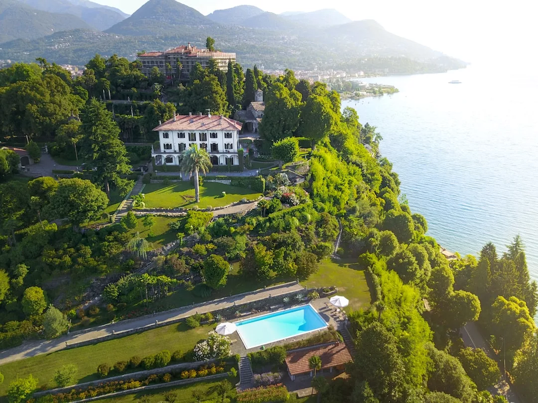 Aerial view of a stunning real estate villa with a swimming pool, nestled among lush greenery on a hillside, overlooking a tranquil lake and majestic mountains in the background.