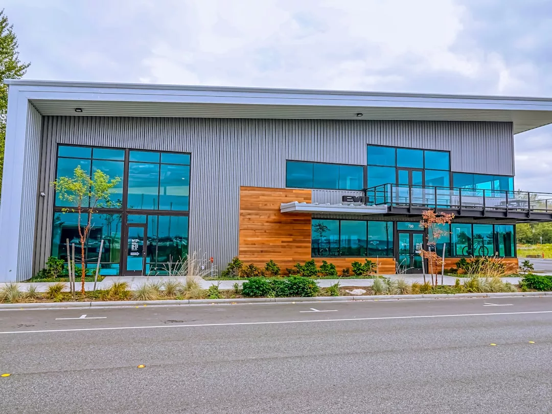 Modern two-story commercial real estate building with large blue-tinted windows, wood accents, metal siding, and a landscaped area in front, viewed from across an empty street.