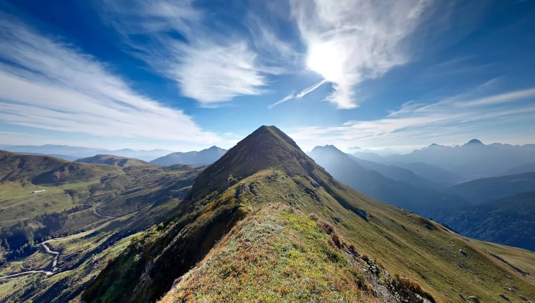 A mountain ridge leads to a peak under a bright sun and blue sky, with clouds and distant mountain ranges visible in the background—a breathtaking view perfect for real estate seekers craving nature’s beauty.