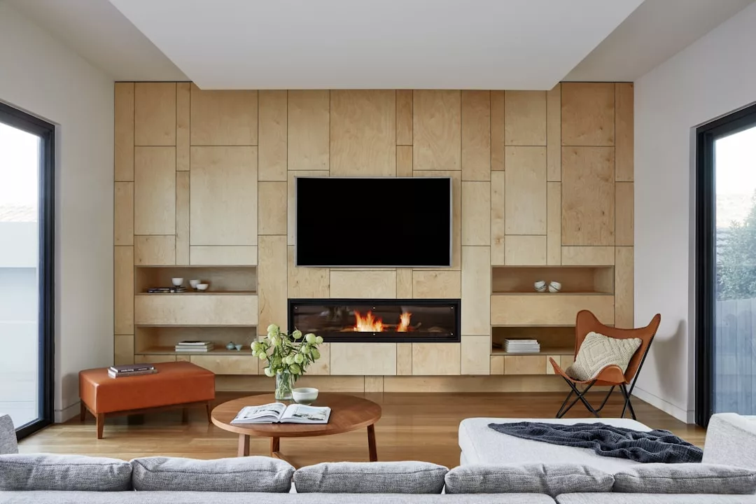 Modern living room with a wood-paneled accent wall, built-in shelves, flat-screen TV, fireplace, gray sofa, round coffee table, orange ottoman, and butterfly chair—a stunning showcase for any real estate listing.