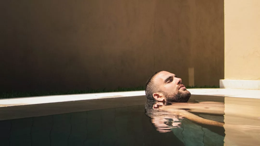 A man relaxes in a pool with his head resting on the edge, eyes closed, bathed in sunlight against a plain beige wall—a serene real estate retreat designed for ultimate comfort and tranquility.