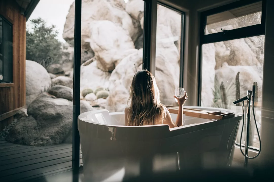 A person with long hair sits in a modern white bathtub by large windows, holding a glass, enjoying a rocky outdoor view—a stunning real estate feature.