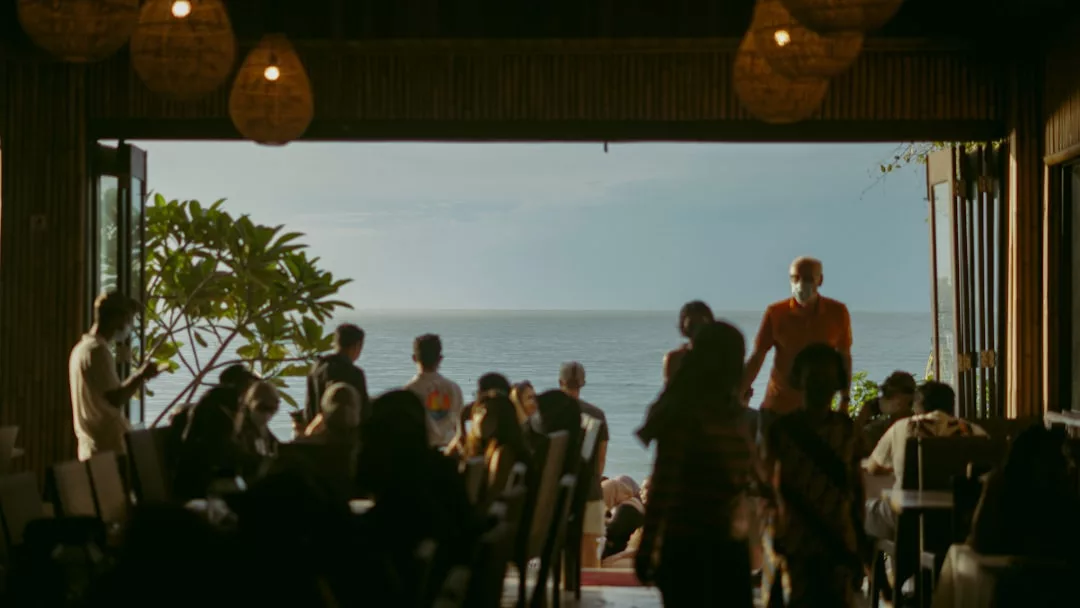 People are seated and standing inside a dimly lit restaurant with open doors, overlooking the sea and a bright sky outside—a scene that highlights the allure of coastal real estate.