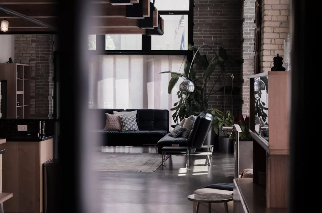 Modern living room with exposed brick walls, large windows, black sofas, a floor lamp, indoor plants, and minimalist decor—perfect for those seeking stylish real estate with contemporary charm.